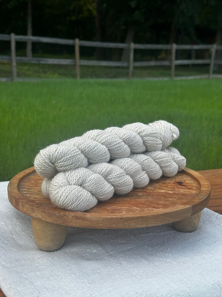 Four skeins of (White Sails) light oatmeal-colored yarn arranged on a round wooden stand outdoors, with a blurred green lawn and fence in the background.